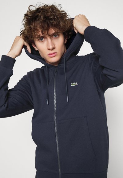 Haut à capuche zippé bleu marine en tissu mélange de coton, avec une poche kangourou, une capuche réglable avec cordon et un petit logo sur la poitrine.