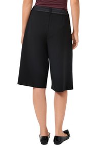 Kobieta ubrana w czarne szorty do połowy uda, czarny pasek, bordową bluzkę i czarne buty na płaskiej podeszwie, pokazana z tyłu na tle białym.