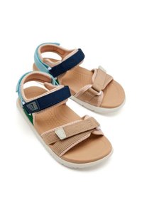 Sandales beiges avec des sangles ajustables en navy, turquoise et vert. Semelle intérieure texturée et semelle extérieure en caoutchouc blanc. Détails de fermeture Velcro.