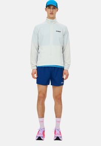 Giacca sportiva leggera bianca e blu con zip abbinata a pantaloni corti blu, sneakers rosa e viola, e calze bianche con dettagli del logo.