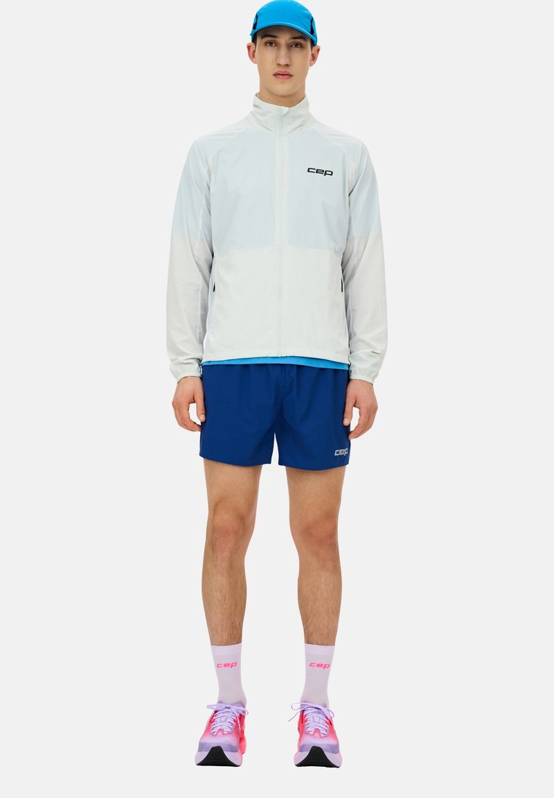 Giacca sportiva leggera bianca e blu con zip abbinata a pantaloni corti blu, sneakers rosa e viola, e calze bianche con dettagli del logo.