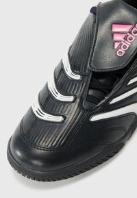 Svart Adidas-sneaker med hvite detaljer, teksturert skinn, rosa logo på tungen, og en tykk gummisåle vinklet for å vise den øvre fronten.