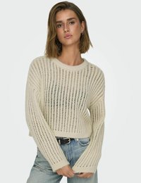Hellbeige gestrickter Pullover mit einem offenen Maschenmuster, langen Ärmeln und einem kurzen Saum, kombiniert mit blauen Jeans.