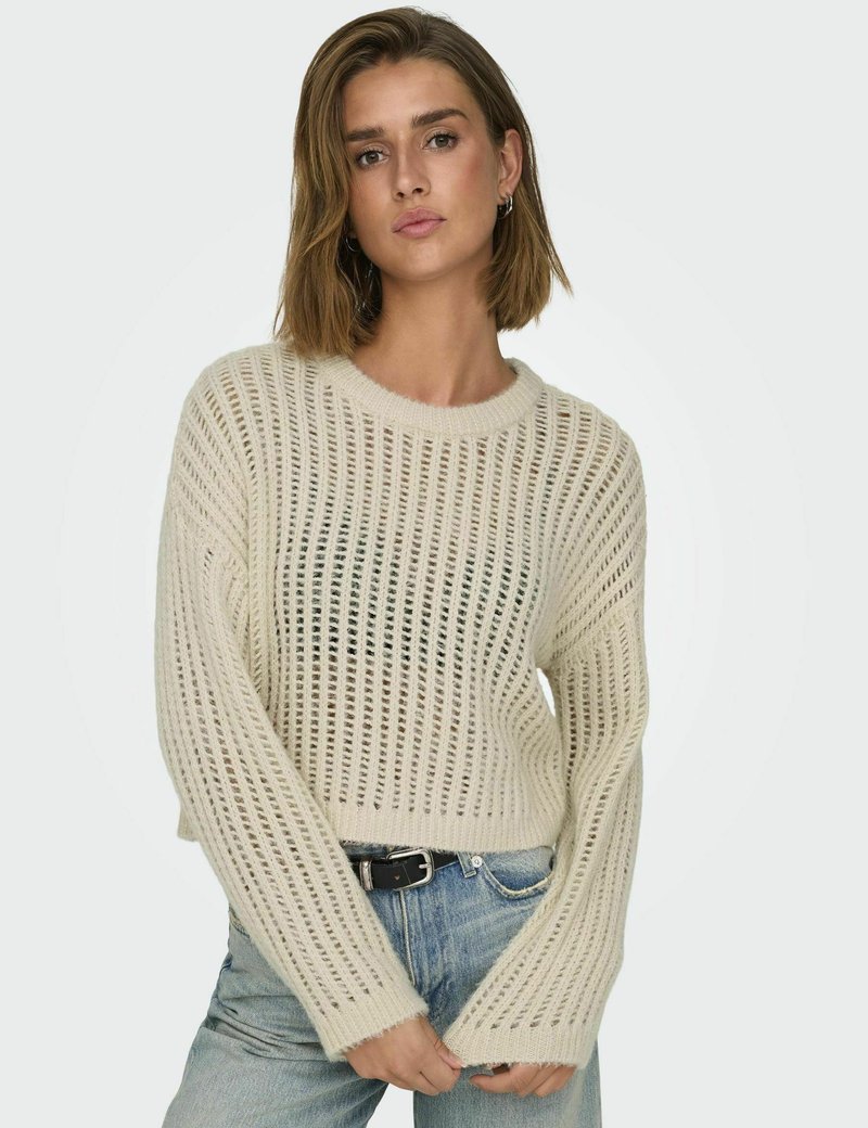 Hellbeige gestrickter Pullover mit einem offenen Maschenmuster, langen Ärmeln und einem kurzen Saum, kombiniert mit blauen Jeans.