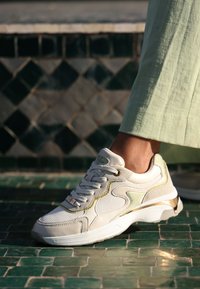 Sneaker bianco e beige chiaro realizzato in materiali in mesh e pelle, con dettagli metallici, suola testurizzata e design con lacci.