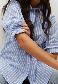 Blue and white striped button-up shirt with rolled sleeves and a relaxed fit. Cotton fabric, subtle sheen, and classic collar design.