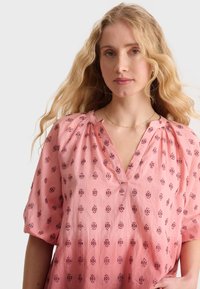 Blusa leggera rosa con scollatura a V, maniche a sbuffo e piccoli motivi floreali in rosa scuro. La texture morbida conferisce un morbido cadente.