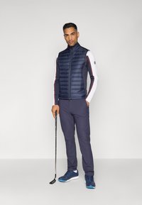 Marineblå puffer vest med vertikal quilting, båret over en hvit langermet skjorte med røde og blå striper. Slim-fit marineblå bukser og blå golfsko.