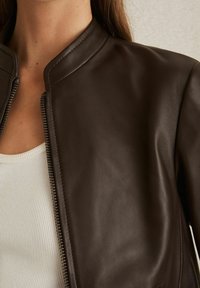 Giacca in pelle marrone con chiusura a zip e collo arrotondato, caratterizzata da una superficie liscia e un design aderente sopra un top bianco a costine.