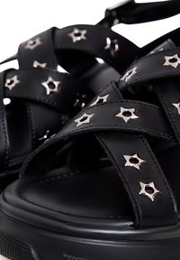Zwarte sandalen met kruislings gespen en metallic steraccenten. Voorzien van een dikke zool en een gladde leren textuur.
