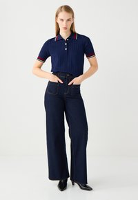 Polo shirt blu navy a coste con colletto a righe rosse e bianche, abbinato a pantaloni a gamba larga in denim scuro a vita alta e scarpe col tacco a punta nere.