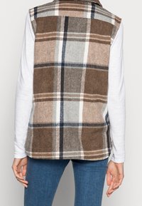 Brun och grårandig ärmlös väst med hög krage, tillverkad av mjuk tyg, lager över en vit långärmad skjorta, kombinerad med blå jeans.