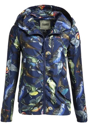 Personne souriante portant une veste à capuche bleu marine à motif d'oiseaux, un jean skinny clair déchiré et des bottes blanches à lacets, debout les mains sur les hanches.