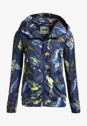 Personne souriante portant une veste à capuche bleu marine à motif d'oiseaux, un jean skinny clair déchiré et des bottes blanches à lacets, debout les mains sur les hanches.