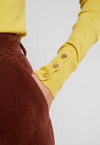 Rukáv žlutého bavlněného svetru s třemi kovovými knoflíky na manžetě, doplněný hnědými manšestráky s žebrovanou strukturou.