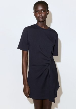 Dark-skinned woman wearing a short-sleeve, knee-length black dress with a side knot detail, standing against a plain white background.