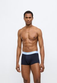 Boxer en coton noir avec une ceinture à logo Lacoste bleu clair, coupe ajustée et texture lisse, modèle posé debout avec le torse nu.