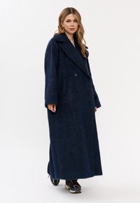 Donna che indossa un lungo cappotto fuzzy blu scuro e sneakers multicolori, in piedi su uno sfondo bianco semplice.