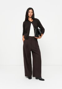 Mørkebrun cardigan med knapper, hvit topp og vide brune bukser. Holder en svart veske, bruker svarte høye hæler. Minimalistisk design.