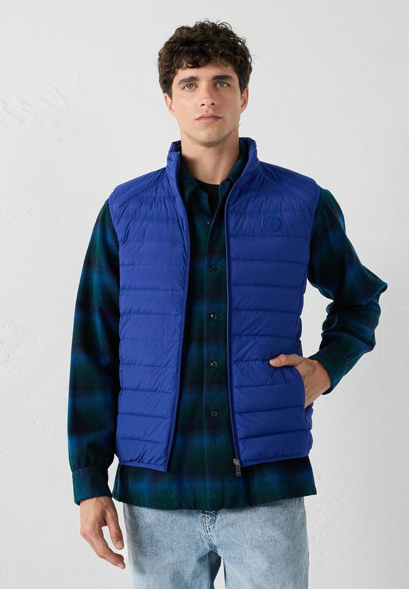 Mann in einer königsblauen Steppweste über einem grün-blauen Karoshirt, der mit einer Hand in der Weste steht, vor einem schlichten Hintergrund.