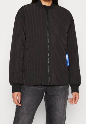 Veste matelassée noire zippée avec poignets côtelés, col et une petite étiquette bleue sur le côté droit, portée par-dessus un jean gris foncé.