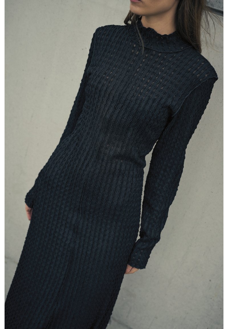 Schwarzes Strickkleid mit langen Ärmeln, das eine gerippte Textur und einen leichten Rollkragen aufweist. Das Design ist figurbetont und bodenlang.