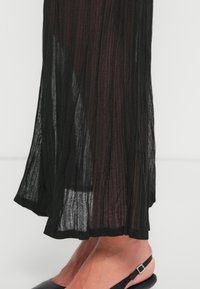 Pieds portant des chaussures slingback noires, partiellement couverts par un tissu noir transparent et texturé d'une longue jupe ou robe.