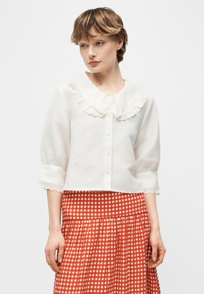 Νεαρός με κοντά μαλλιά φορώντας ένα λευκό μπλουζάκι με βολάν στο γιακά και τα μανίκια, συνδυασμένο με μια κόκκινη και λευκή καρό φούστα.