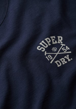Granatowy materiał z białym nadrukiem tekstu "SUPER DRY." oraz "1954" przecinanym przez dwa kije polo z małym logo na środku.