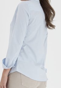 Jasnoniebieska koszula zapinana na guziki o gładkiej fakturze, z długimi rękawami podwiniętymi przy mankietach i zaokrąglonym dołem. Nosi się ją z beżowymi spodniami.