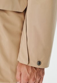Beżowy rękaw kurtki z gładką teksturą, z bocznym rozcięciem oraz detalem guzika na mankiecie z metalicznym akcentem.