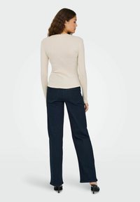 Langermet kremfarget topp kombinert med mørkeblå vide jeans, med en glatt tekstur og subtile sømdetaljer, plassert på en ensfarget bakgrunn.