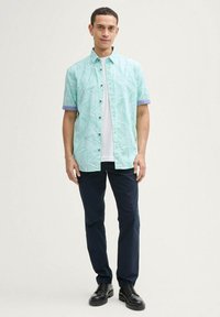 Korte mouwen shirt in licht mintkleurige stof met een bladerenpatroon, gecombineerd met marineblauwe broek en zwarte schoenen. Achtergrond is eenvoudig.