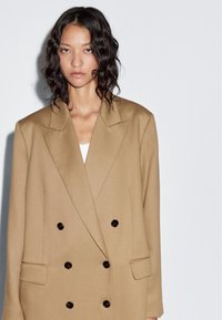 Double-breasted blazer in beige fabric with structured shoulders, large black buttons, and notch lapels. Features side pockets and a relaxed fit.
