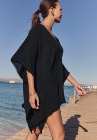 Črni teksturiran pončo s cofki, z rahlo prilegajočim in V-izrezom, ki ga nosi oseba na lesenem pomolu ob vodi.