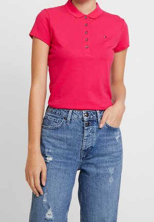 Kvinde iført en tætsiddende rød poloshirt med korte ærmer og knapper, kombineret med højttaljede, slidte blå jeans, med den ene hånd i lommen.