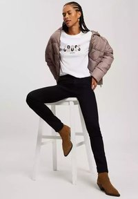 Puchowa kurtka w jasnobrązowym kolorze, biała koszulka z napisem "CROSS JEANS", czarne dopasowane spodnie oraz brązowe botki za kostkę z ostrymi noskami.