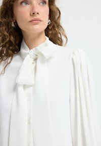 Hvid bluse med høj hals og sløjfe-detalje. Har lange, samlet ærmer og en glat stoftekstur. Knaplukning foran.