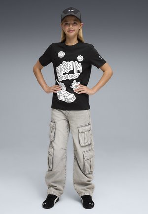 Une fille se tient les mains sur les hanches, portant un t-shirt noir BMW Motorsport, un pantalon cargo gris, des chaussures noires Puma et une casquette BMW, sur un fond gris.