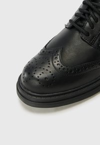 Sorte skinnsko med dekorative perforeringer og søm. Har en rund tå og flat gummisåle. Klassisk wingtip-design.