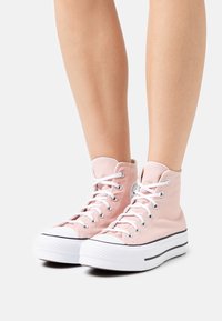 Zapatillas altas de lona suave en rosa pastel con cordones blancos y suelas de goma. Presentan un logo circular y ribete negro contrastante.
