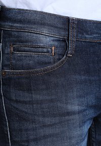 Tmavomodré denimové džínsy s klasickým dizajnom, s viditeľným prešívaním a malým detailom predného vrecka. Textúrovaná tkanina s jemným leskom.