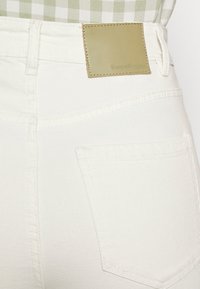 Calças de ganga brancas com uma superfície texturizada, bolso traseiro e um retângulo de pele verde rotulado com "Denim Project" na cintura.