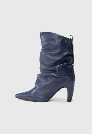 Bota de couro azul-marinho, com bico afiado e design franzido, e um salto alto. Apresenta um ligeiro brilho e uma alça na parte traseira.