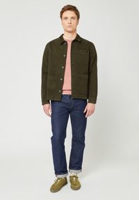 Olive green, button-up jacket with two front pockets, worn over a pink sweater, paired with dark blue jeans and green sneakers.