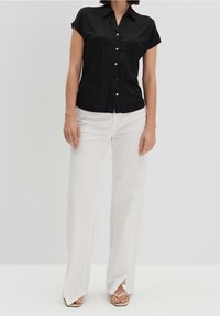 Camisa negra de manga corta y botón, hecha de un material suave, combinada con pantalones blancos de pierna amplia con aberturas laterales y zapatos de sandalia de color claro.