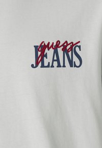 Balta medvilninė marškinėlių su raudonai siuvinėtu užrašu "guess" ir tamsiai mėlynu, paryškintu žodžiu "JEANS". Lygus paviršius, kasdienis dizainas.