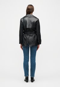 Γυναίκα με μακριά καστανά μαλλιά που  φοράει μαύρο δερμάτινο μπουφάν με ζώνη, μπλε τζιν και μαύρες μπότες μέχρι τον αστράγαλο, όρθια με την πλάτη προς το μέρος μας σε λευκό φόντο.