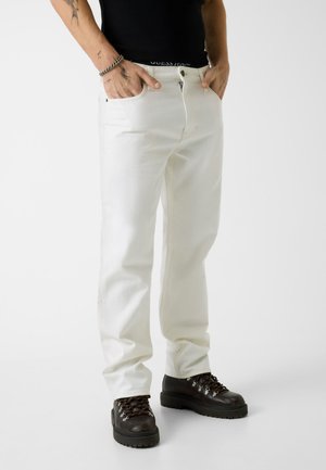 Person, die weiße Jeans trägt, die Hände in den Taschen hat, ein schwarzes Oberteil und dunkelbraune Schnürstiefel trägt und auf einem schlichten weißen Hintergrund steht.