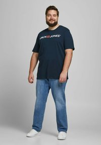 Marineblaue Baumwoll-T-Shirt mit dem "JACK & JONES"-Logo, kombiniert mit hellblauen Jeans und weißen Sneakers, vor einem grauen Hintergrund stehend.